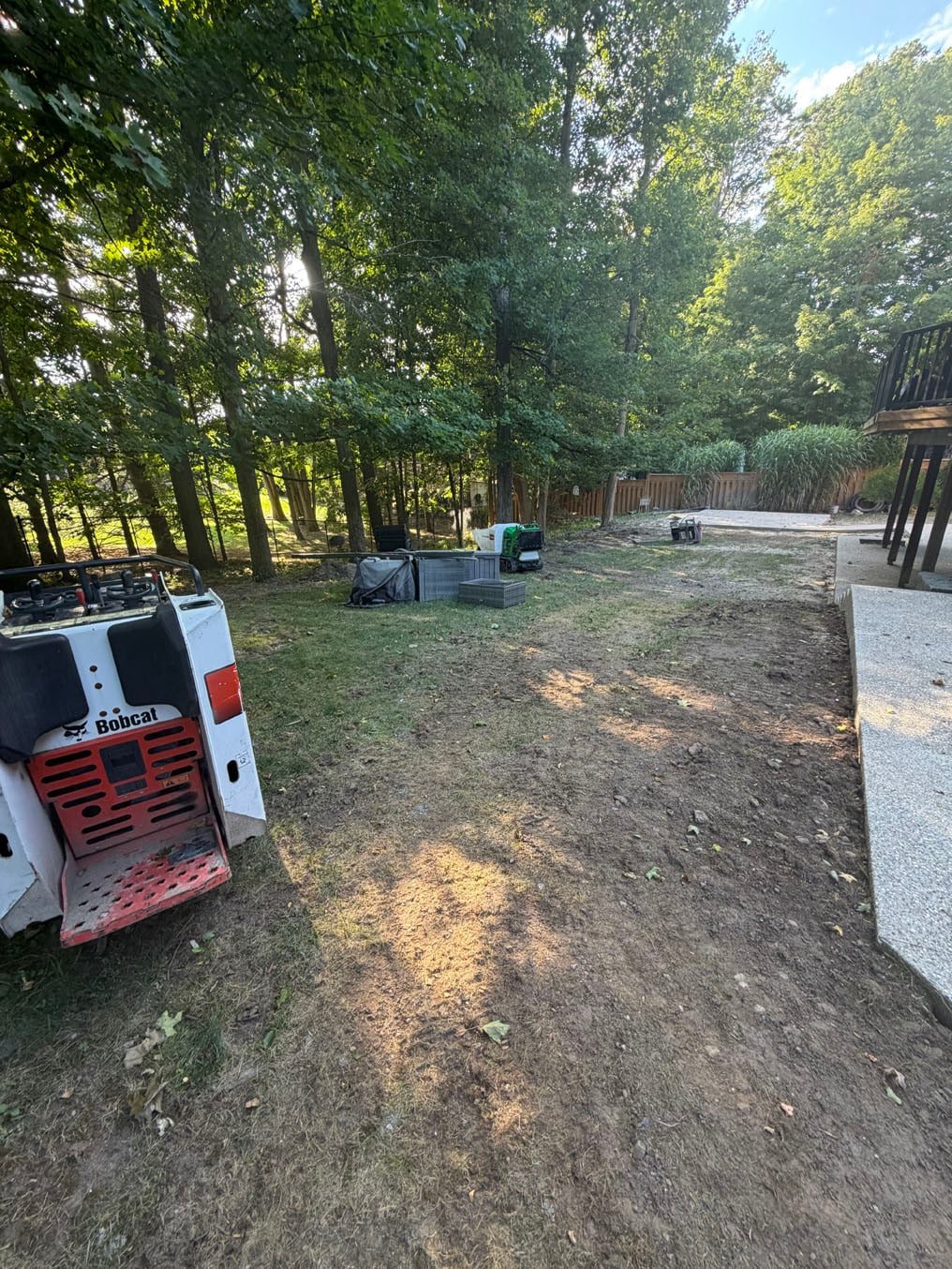 Sod Installation On Carrington Place, Waterloo 1 Bare, patchy back yard under tall trees with a skid steer on site behind a Carrington Place, Waterloo home