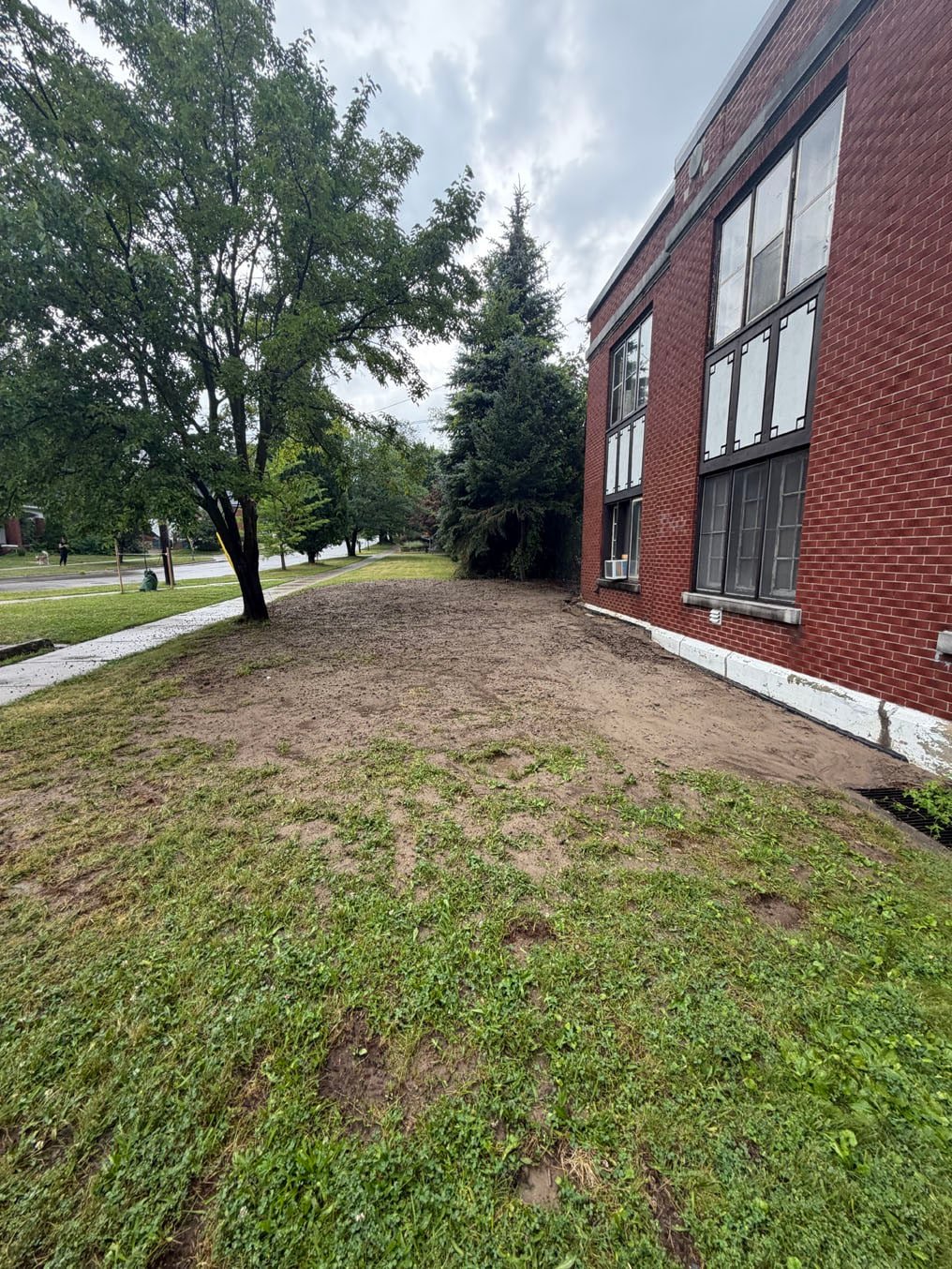 Bare, weedy ground along the side of a two-storey brick building in Cherry Hill, Kitchener, before sod installation