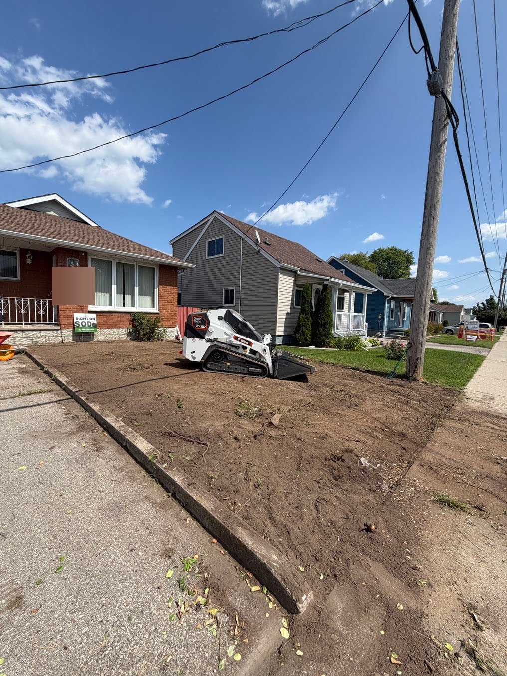 Front yard in Galt, Cambridge cleared to bare soil from the driveway curb to the sidewalk, the track loader parked on it and the boulder gone