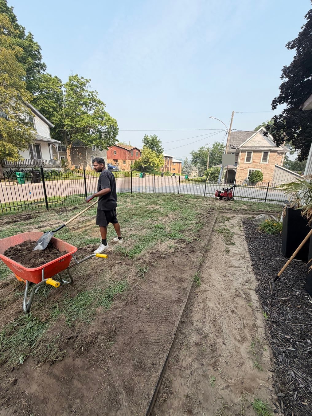 Crew member shovelling old turf into a wheelbarrow beside the garden bed at the porch in Hespeler, Cambridge, sod cutter parked at the iron fence