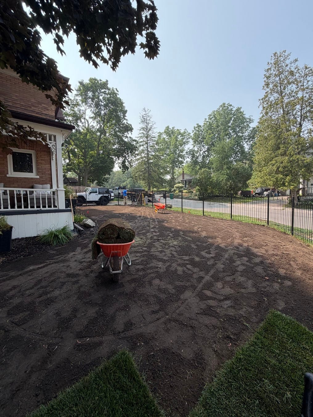 Fresh topsoil graded across the fenced front lawn in Hespeler, Cambridge, first sod rolls down at the edges and a wheelbarrow of rolls near the porch