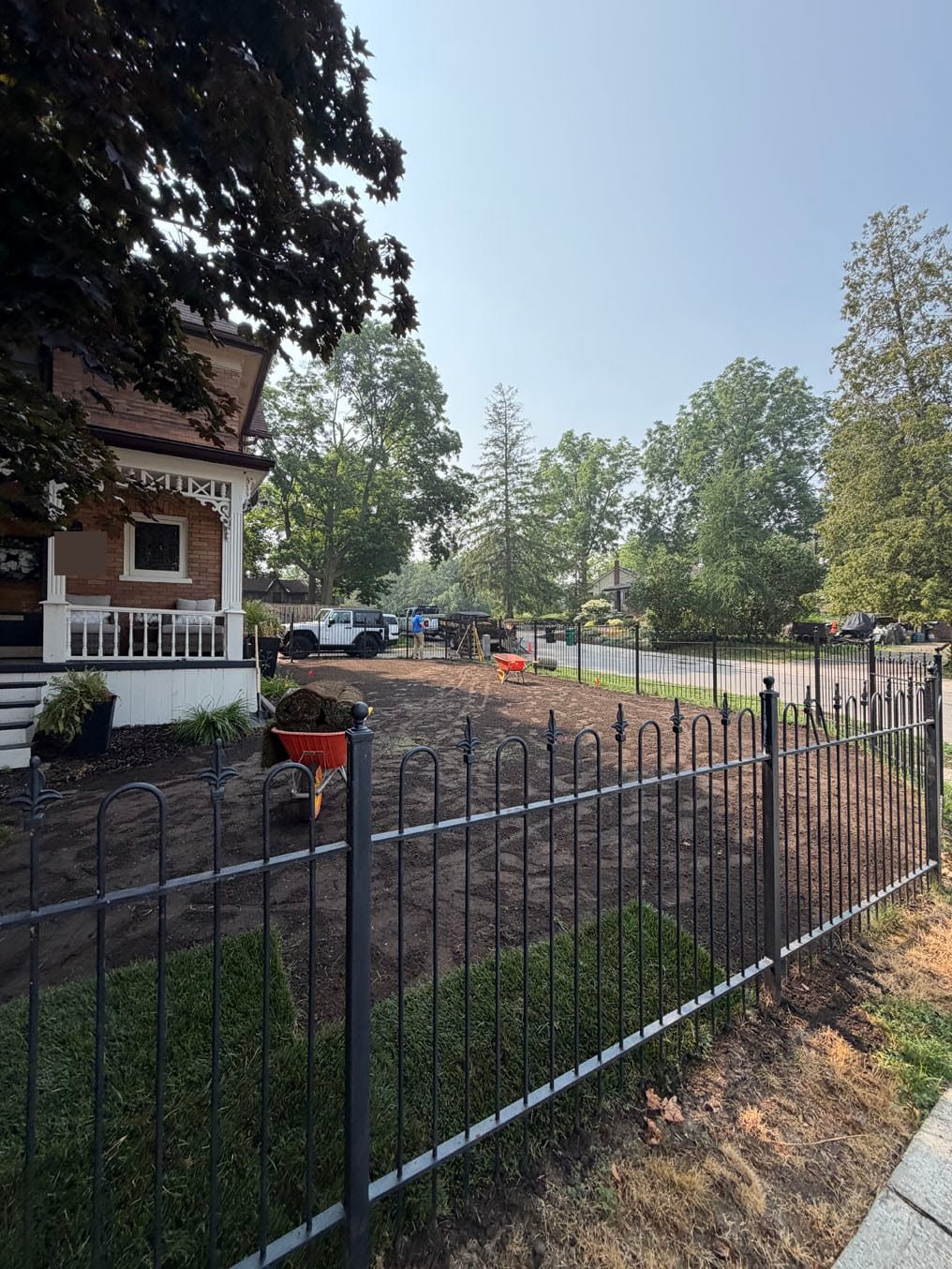 Sod rows laid along the iron fence at the brick home in Hespeler, Cambridge, graded topsoil and a wheelbarrow of rolls behind
