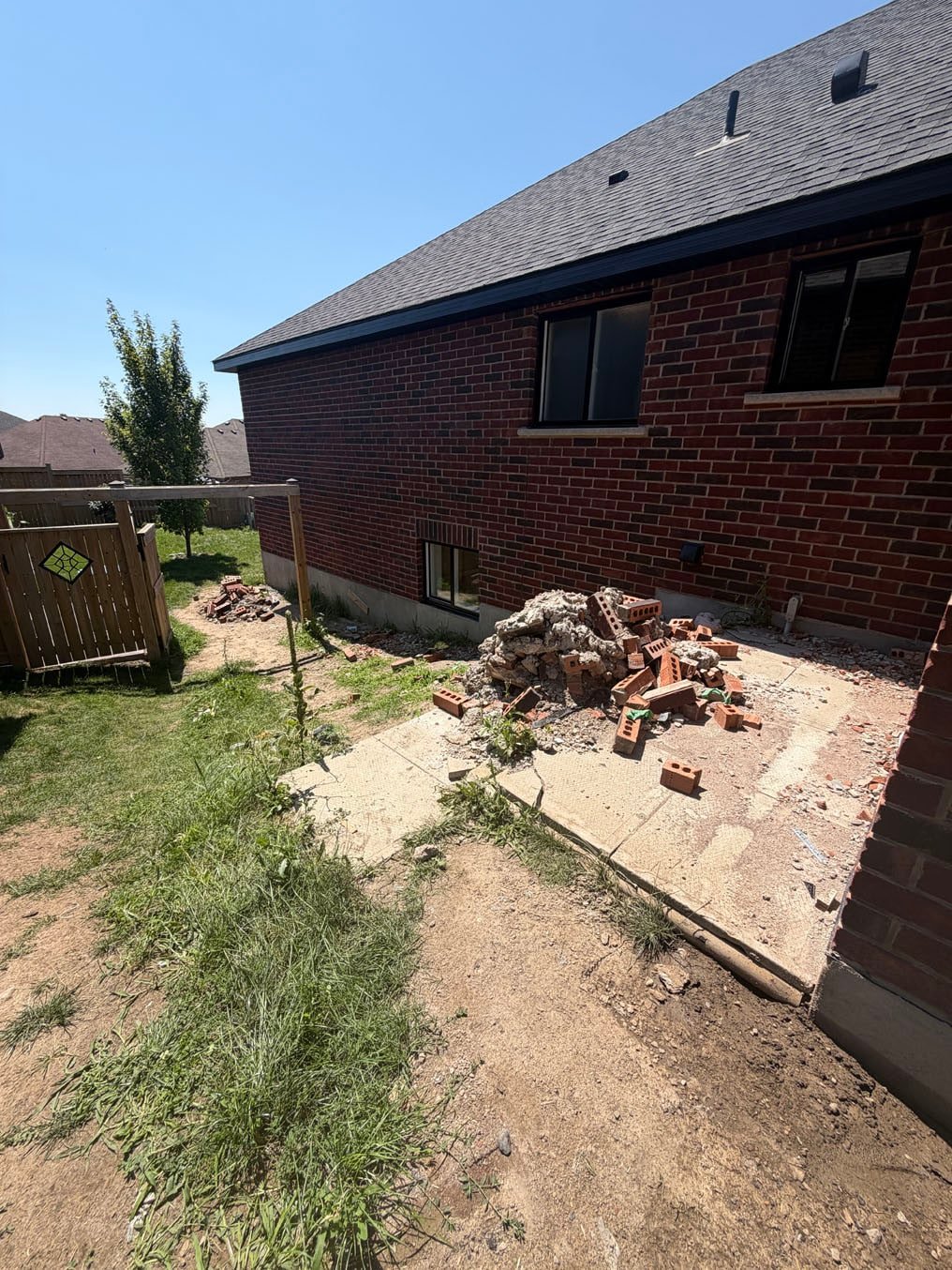Worn, weedy side yard with a bare dirt path through to the backyard beside a red-brick bungalow in Plattsville, Ontario, before sod