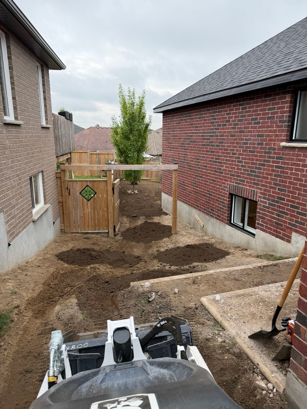 Sod Installation In Plattsville, Ontario 8 Fresh topsoil piles dumped along the stripped side yard, seen from the skid steer with the arbor ahead, Plattsville, Ontario