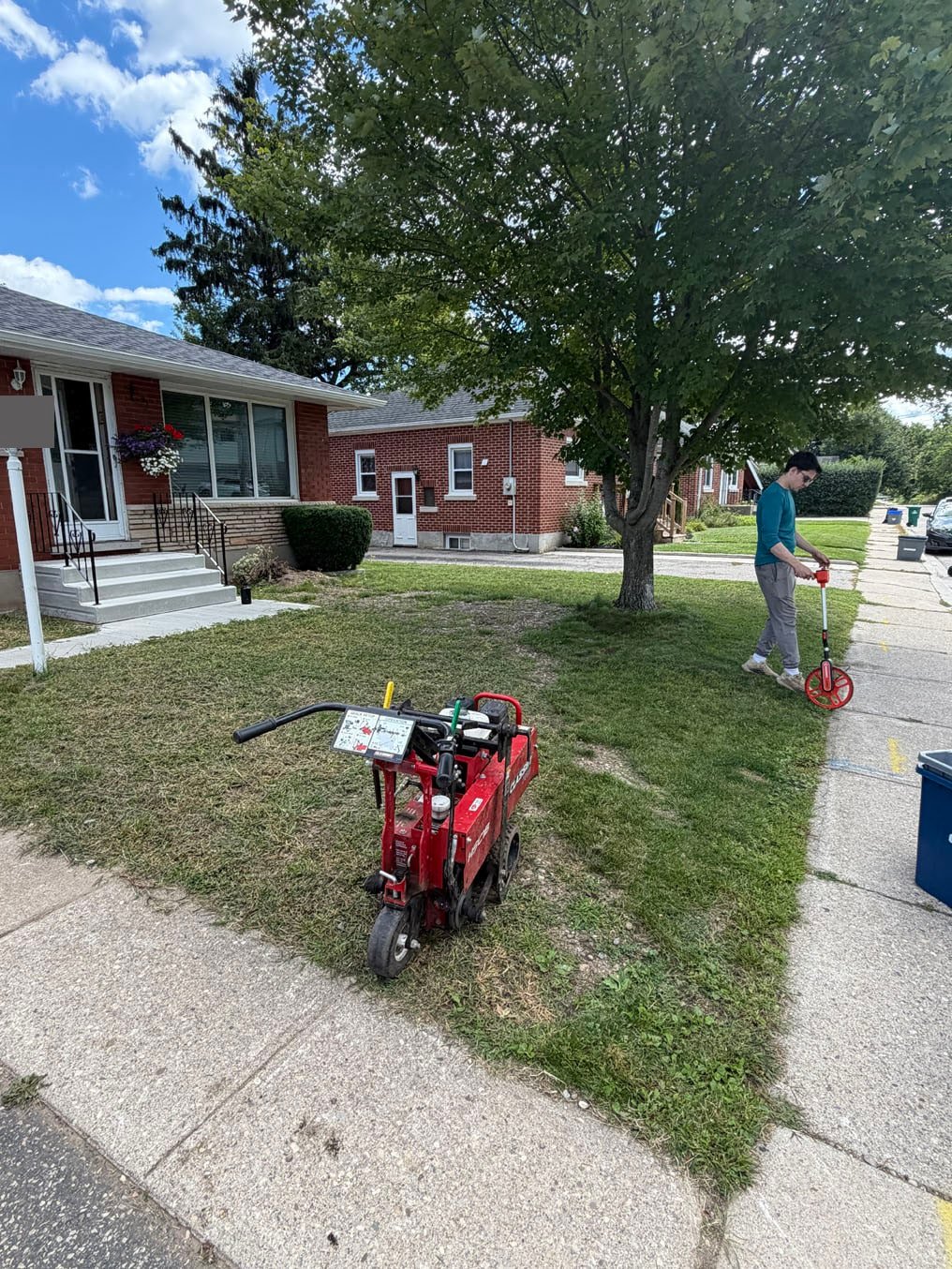 Patchy, worn front lawn at a red-brick bungalow in Preston, Cambridge, sod cutter on the grass and a crew member measuring it before sod replacement