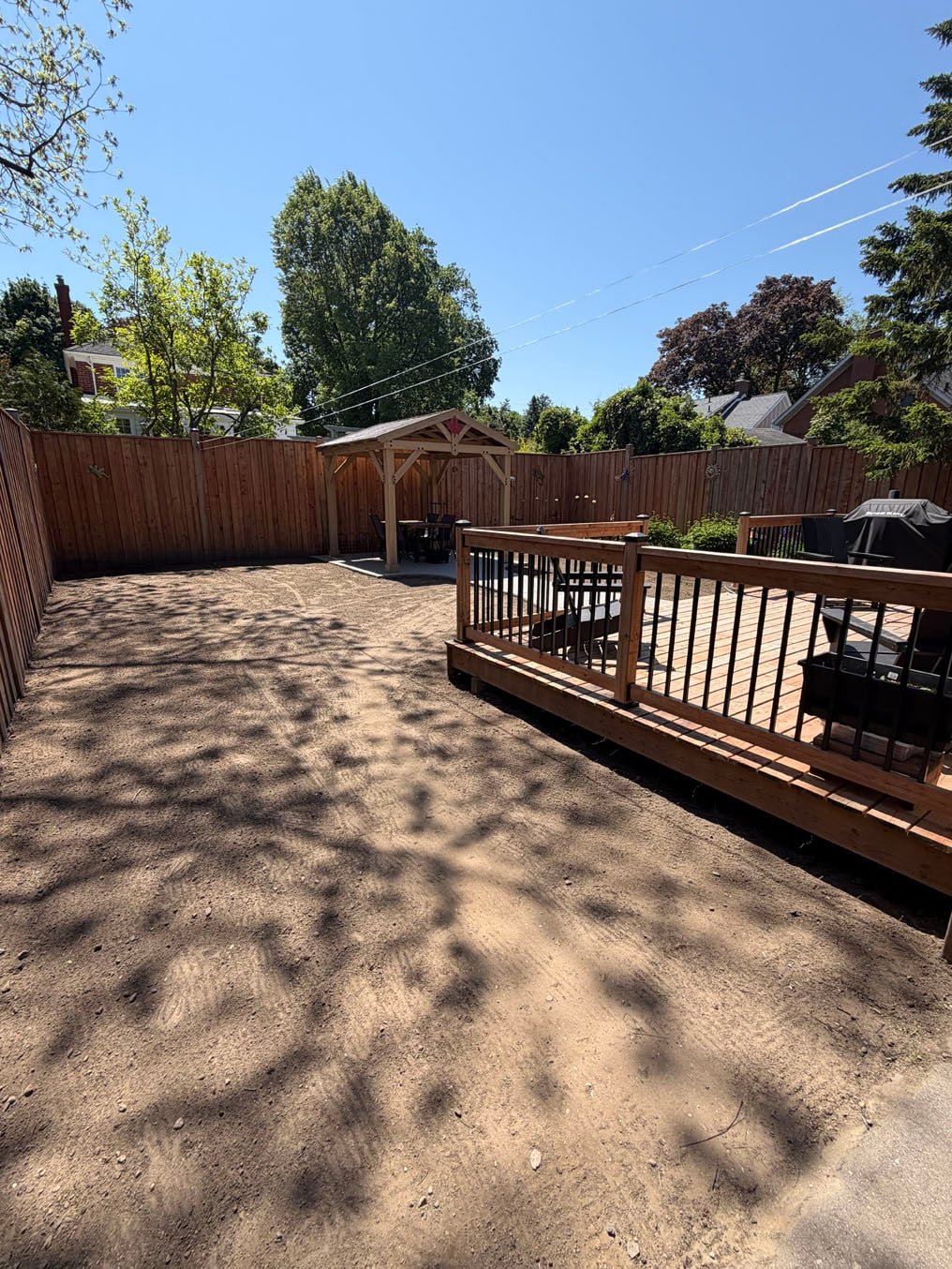 Bare, packed ground between a new deck and a gazebo in a fenced back yard in Westmount, Waterloo, before sod